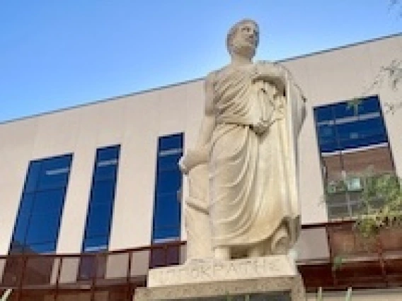 A white statue of Hippocrates stands in front of a tan building with tall windows.