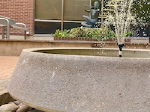 A concrete circular fountain sprays water in front of a brick building with large windows.
