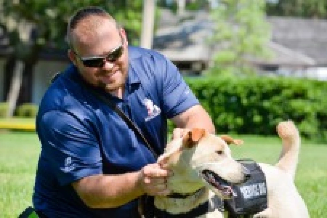 A man wearing sunglasses and a navy blue polo shirtkneels next to a medium-sized yellow dog wearing a "Service Dog" vest.