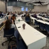Attendees fill a large lecture hall during the July 30 Conflict Resolution and De-escalation Skills Symposium at the University of Arizona. Participants sit in groups at long white tables, engaging in discussion during an interactive portion of the event. Multiple presentation screens display a slide with the word “practice” and a blue circular graphic divided into four sections.