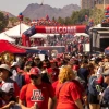 A large crowd of University of Arizona fans fills the University of Arizona Mall during Homecoming festivities, surrounded by red and blue tents, balloons, and a “Welcome” archway with mountains visible in the background.