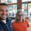 Josh Wright and Jordan Orvik smile for a photo inside a café with large windows. Josh, wearing a navy hoodie, stands on the left with his arm around Jordan, who is wearing glasses and an orange ‘Class of 2022’ T-shirt. Outdoor tables and people sitting on the patio are visible through the windows behind them.